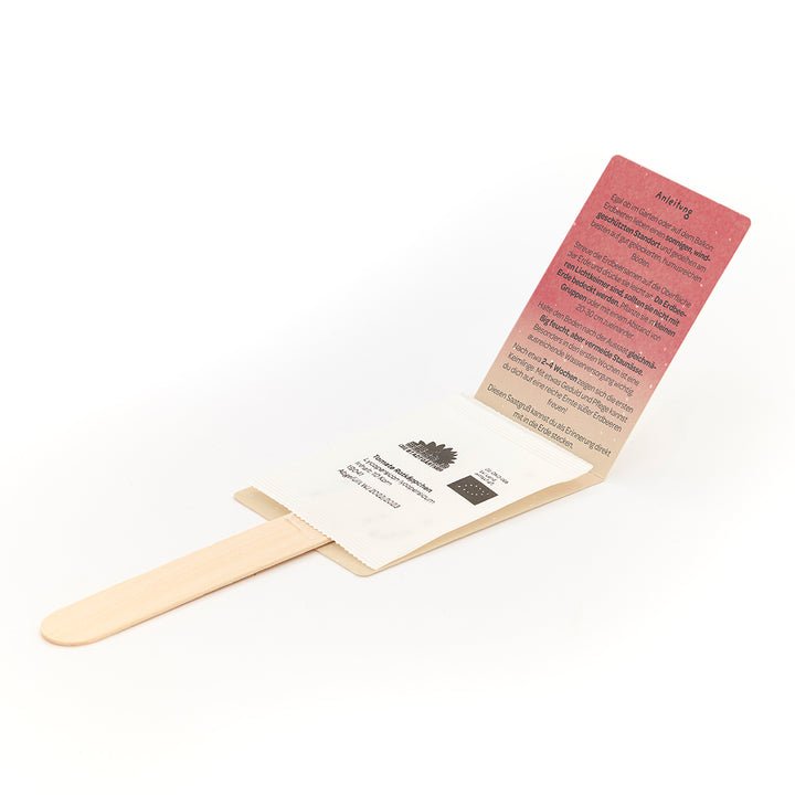 Ein Saatgutset in Form eines Holzstiels mit aufgeklappter Papierverpackung und Anweisungen auf Deutsch.
