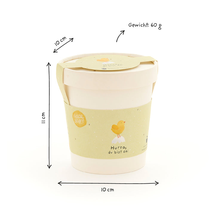 Beiger Becher mit gelbem Etikett, Küken-Illustration und Text "Hurra, du bist da!", mit Maßangaben (10 cm Durchmesser, 11 cm Höhe) und Gewicht (60 g) auf Deutsch.