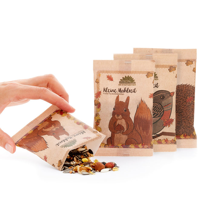 "Hand öffnet eine kleine Papiertüte mit Tierfutter neben zwei weiteren Tüten mit Eichhörnchen- und Igelillustrationen"