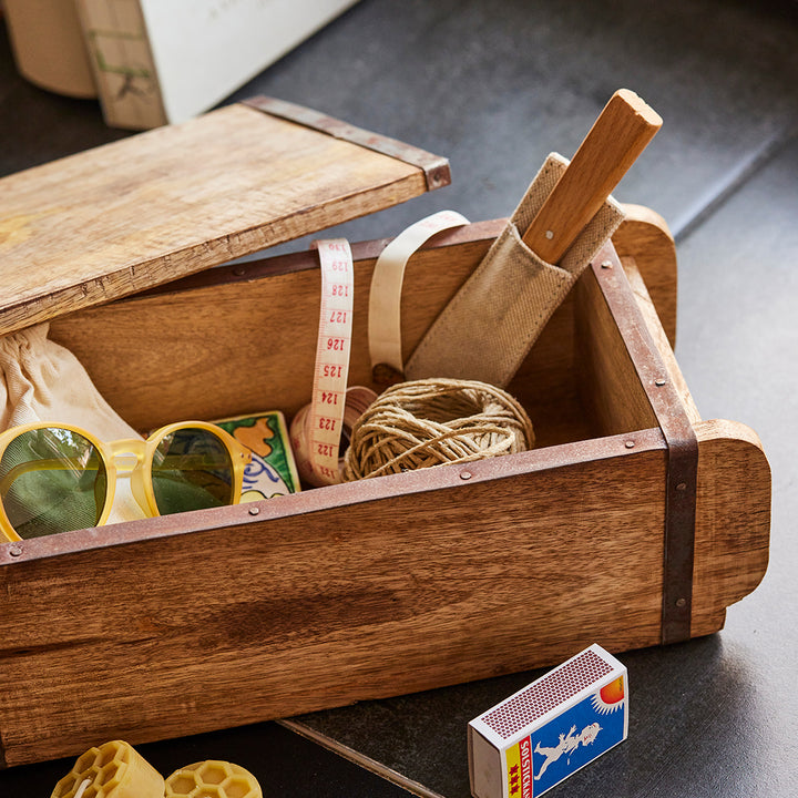 Holzkiste mit verschiedenen Gegenständen, darunter eine gelbe Sonnenbrille, ein Maßband, eine Rolle Schnur, eine Schachtel Streichhölzer und ein Holzgriffwerkzeug.