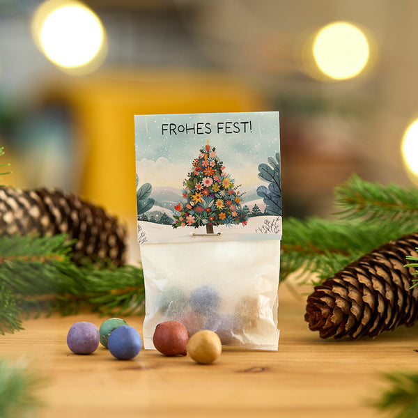 Tüte mit bunten Kugeln vor einer Karte mit Weihnachtsbaum und Schriftzug "Frohes Fest", umgeben von Tannenzweigen und einem Tannenzapfen.