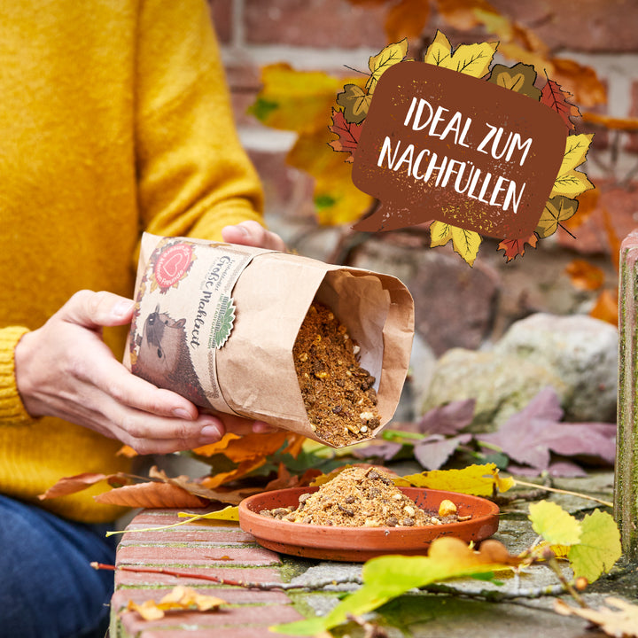Eine Person in gelbem Pullover füllt Igel-Futter aus einer Papiertüte in eine flache Schale, umgeben von Herbstlaub; daneben ein Schild mit der Aufschrift "Ideal zum Nachfüllen".