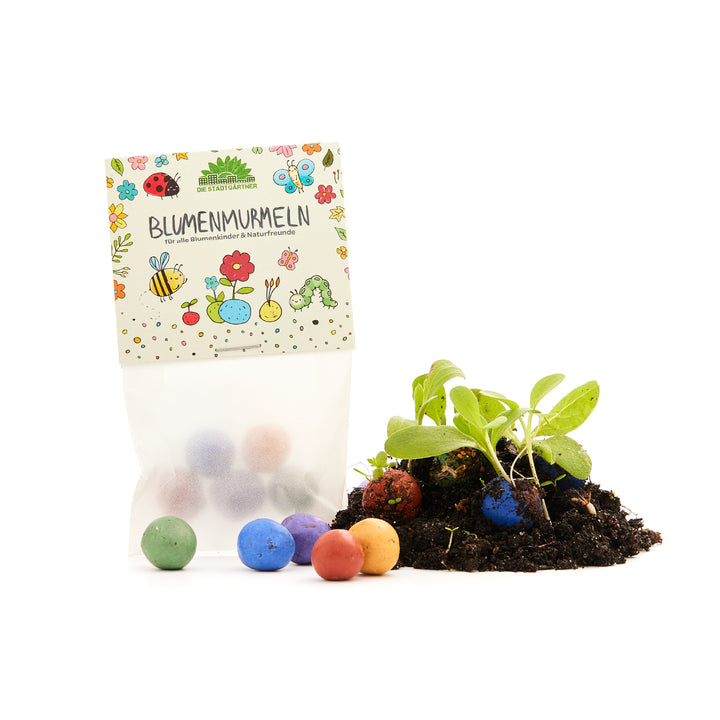 Tüte mit bunten Blumenmurmeln neben Erde, aus der kleine Pflänzchen wachsen.