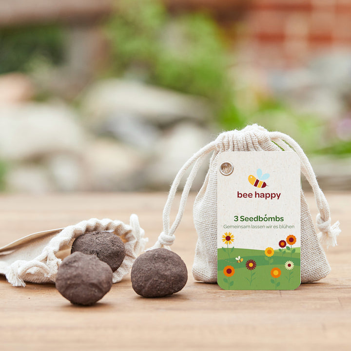 Samenbomben neben einem Beutel mit der Aufschrift "bee happy 3 Seedbombs" auf einem Holztisch mit unscharfem grünem Hintergrund.