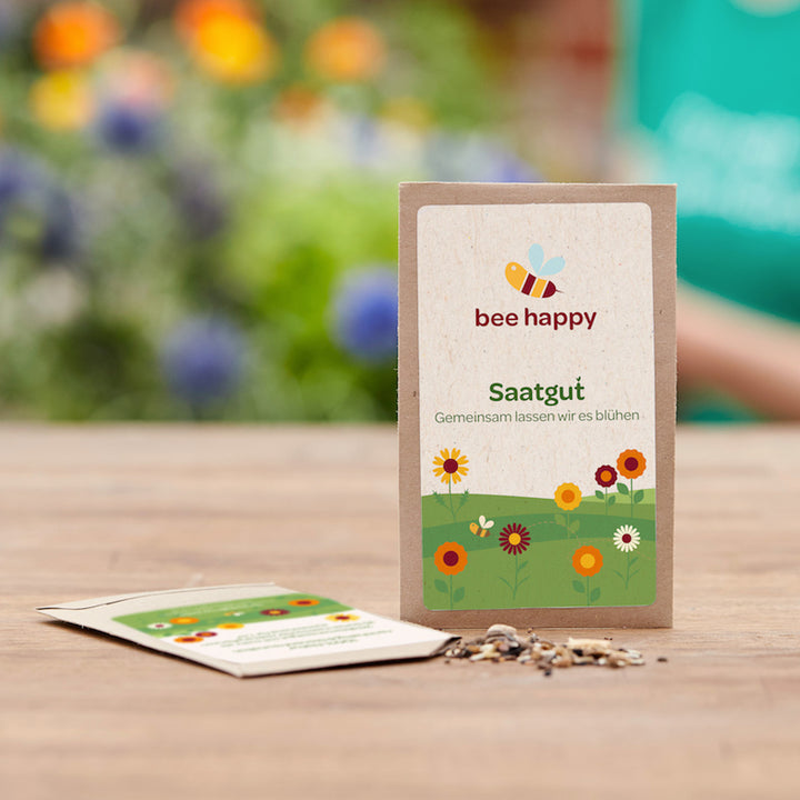 Tüte mit Blumensaatgut „bee happy“ auf einem Holztisch, im Hintergrund unscharfe Blumen.