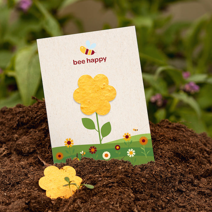 Eine Karte mit einer gelben Blume und der Aufschrift „bee happy“ steckt in Erde, im Vordergrund liegt eine gelbe Blume aus Papier; im Hintergrund grüne Pflanzen.