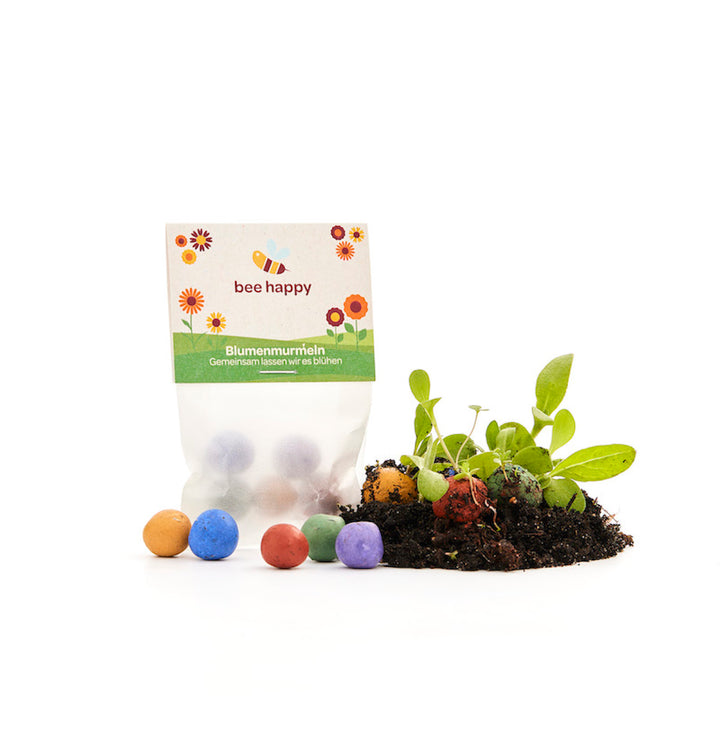 Eine Packung "bee happy" Blumenmurmeln mit bunten Samenbällchen und kleinen Pflanzen auf Erde.