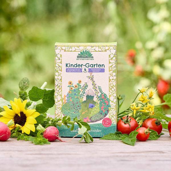 Eine Packung „Kinder-Garten“ Bio-Saatgut steht auf einem Holztisch, umgeben von Sonnenblume, Radieschen, Tomaten und frischem Grün im Garten.