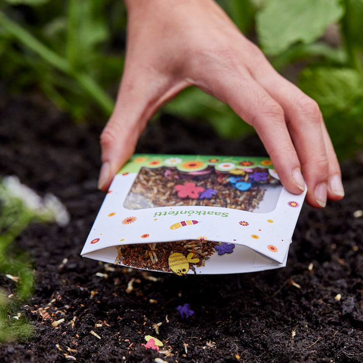 Eine Hand streut Blumensamen aus einer bunt verzierten Tüte auf Erde im Garten.