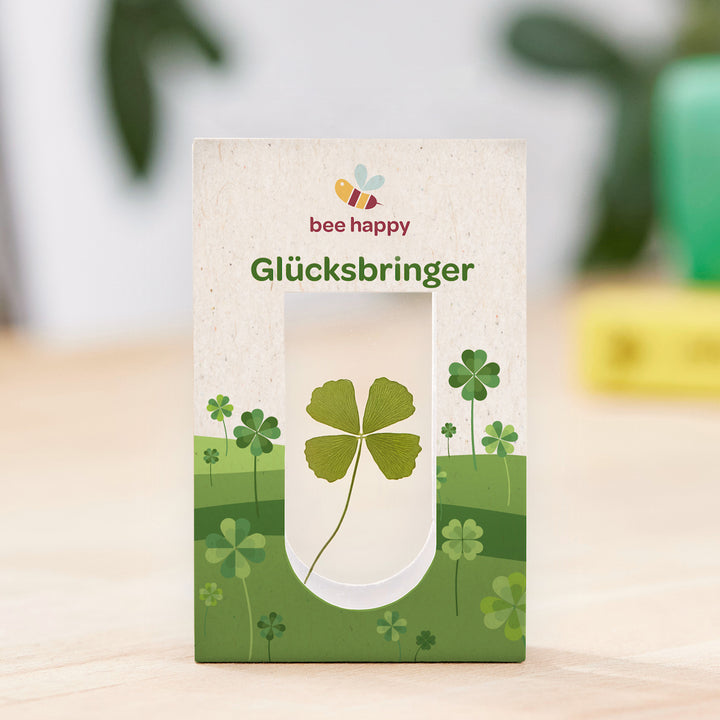 Glücksklee in Geschenkverpackung mit der Aufschrift "bee happy Glücksbringer" auf einem Holztisch.