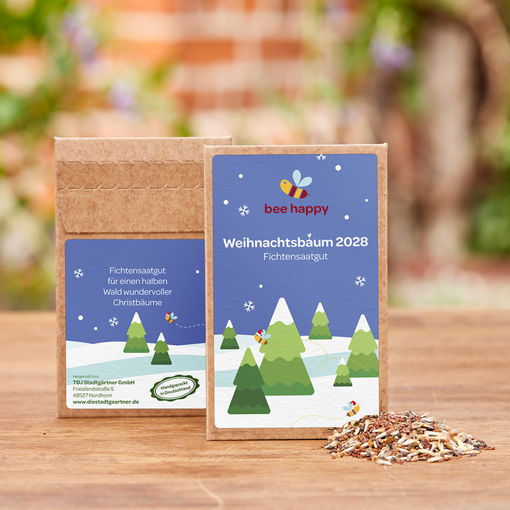 Zwei Packungen mit Fichtensaatgut „Weihnachtsbäum 2028“ auf einem Holztisch, daneben eine kleine Menge Saatgut, im Hintergrund unscharfe Natur.