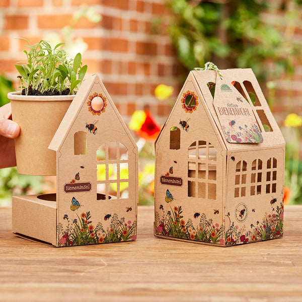 Zwei dekorative, kartonierte Häuschen mit Blumen- und Insektenmotiven, eines mit einem Pflanztopf mit grünen Keimlingen, beide auf einem Holztisch im Garten platziert.
