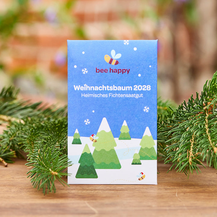 Packung mit der Aufschrift „Weihnachtsbaum 2028 Heimisches Fichtensaatgut“ und buntem Bienen- und Tannenbaum-Design, dekoriert mit Tannenzweigen im Vordergrund.