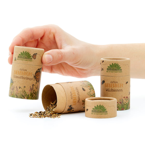 Hand öffnet eine Kartonröhre mit Aufschrift 'Aktion BIENENWOHL' und Samen für Wildbienen und Schmetterlinge.