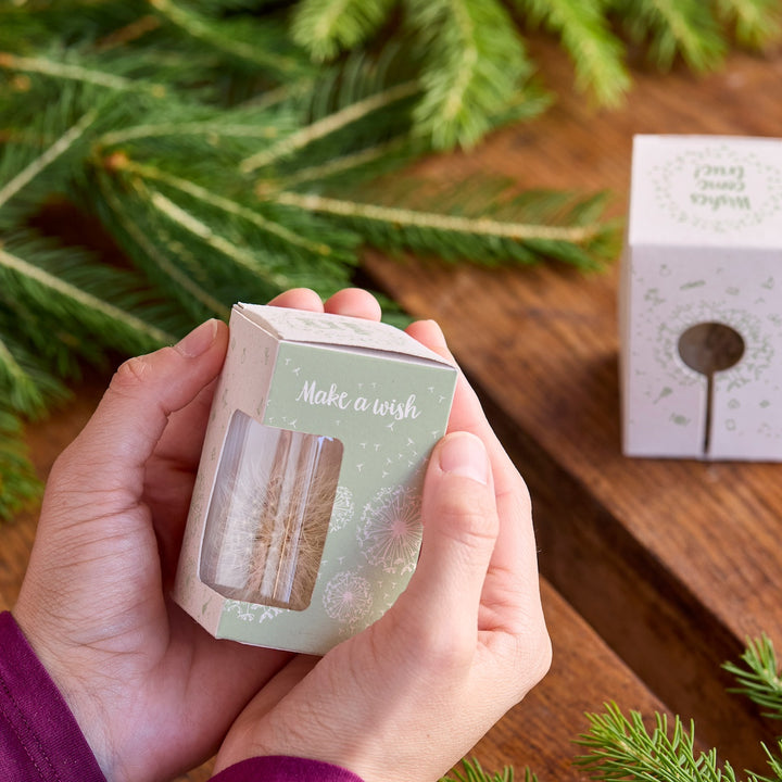 Hände halten eine kleine Geschenkbox mit der Aufschrift "Make a wish" neben Tannenzweigen auf einem Holztisch.