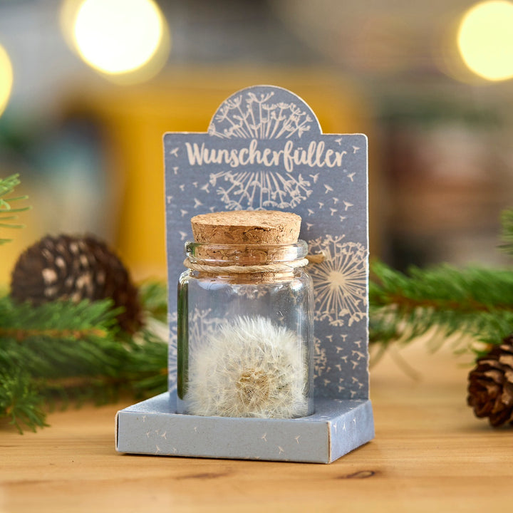 Glasfläschchen mit Löwenzahnpusteblume und Korken, auf einer dekorativen Verpackung mit der Aufschrift „Wunscherfüller“, im Hintergrund Tannenzweige und Zapfen.