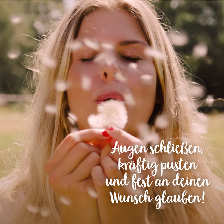 Junge Frau pustet auf eine Pusteblume mit schließenden Augen in einem sonnigen Feld.