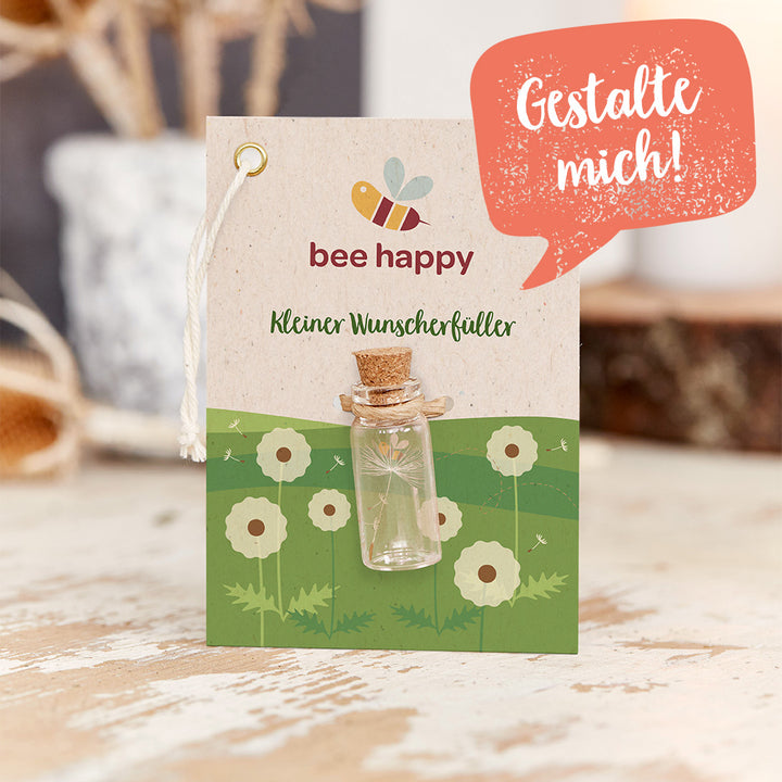 Karte mit der Aufschrift "bee happy" und "Kleiner Wunscherfüller", darauf befestigtes kleines Glasfläschchen, Hintergrund mit gezeichneter Wiese und Pusteblumen, Sprechblase mit dem Text "Gestalte mich!".
