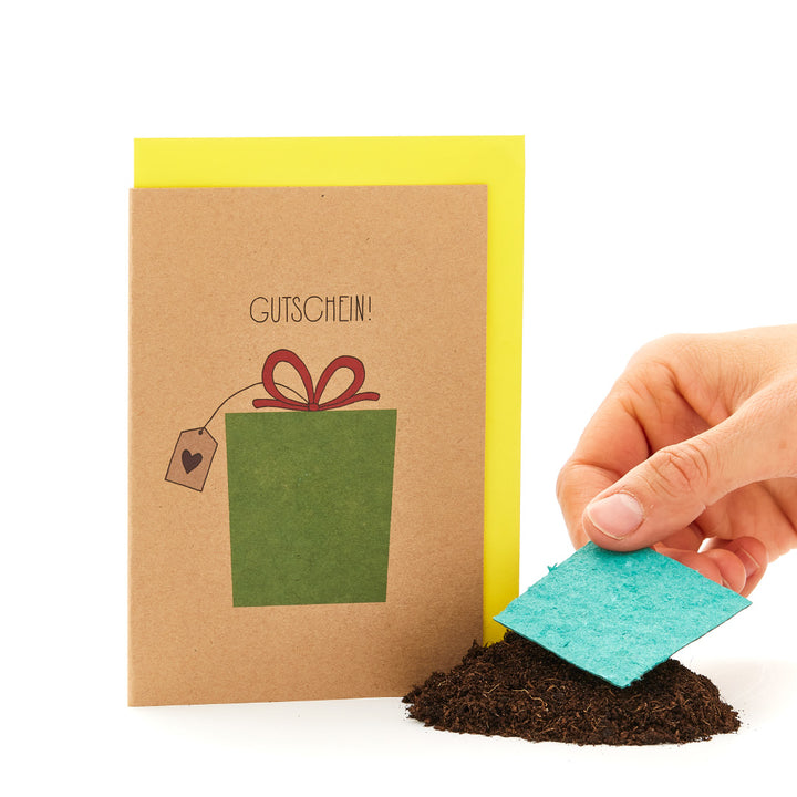 Hand hält eine Grußkarte mit der Aufschrift "GUTSCHEIN" und einem gezeichneten Geschenk, neben einem Haufen Erde mit einem Stück blauer Pappe.