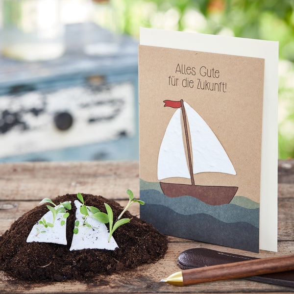 Grußkarte mit Segelboot und der Aufschrift "Alles Gute für die Zukunft!", daneben Erde mit Keimlingen und ein Stift auf einem Holztisch.