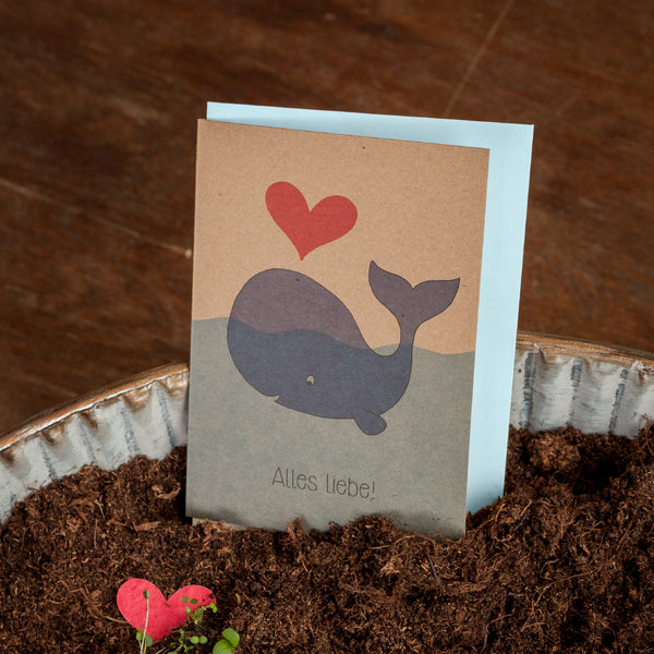 Glückwunschkarte mit einem gezeichneten Wal, einem roten Herz und dem Text "Alles Liebe!" auf Erde stehend.