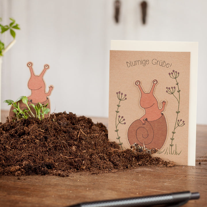 Grußkarte mit Schneckenmotiv und der Aufschrift "blumige Grüße" neben einem Haufen Erde auf einem Holztisch.