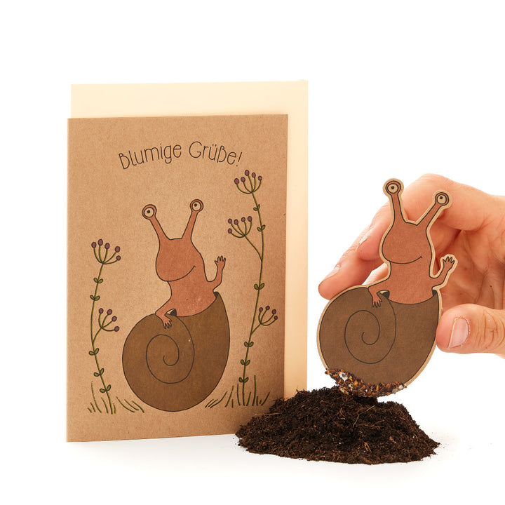 Person hält eine ausgeschnittene Papierillustration einer Schnecke über echter Erde, passend zu Grußkarte mit Text "Blumige Grüße!" und Schneckenzeichnung.