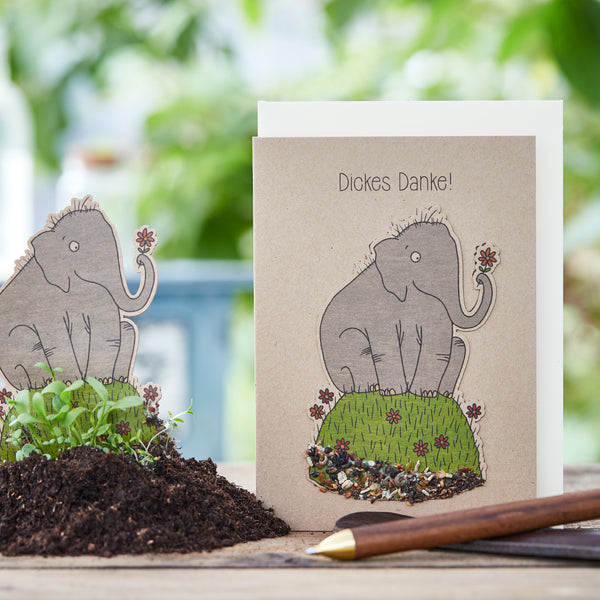 Grußkarte mit gezeichnetem Mammut, auf der „Dickes Danke!“ steht, daneben Erde und grüne Keimlinge.