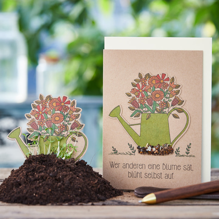 Grußkarte mit einer Illustration einer Gießkanne voller Blumen und dem deutschen Spruch „Wer anderen eine Blume sät, blüht selbst auf“, daneben eine Aussaatkarte im Blumentopf mit Erde und aufgehendem Grün.