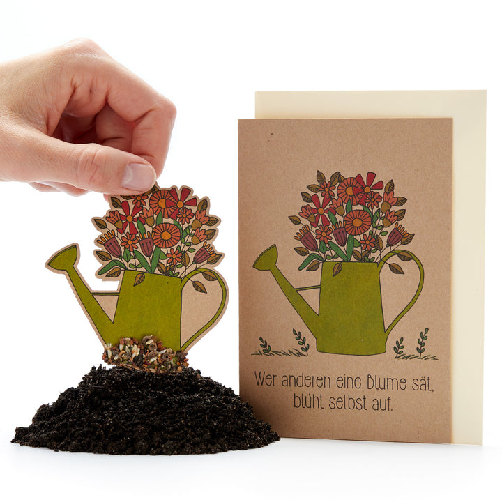 Eine Hand steckt eine mit Blumen verzierte Gießkanne aus Papier in Erde; daneben steht eine Karte mit dem gleichen Motiv und dem deutschen Spruch: "Wer anderen eine Blume sät, blüht selbst auf."