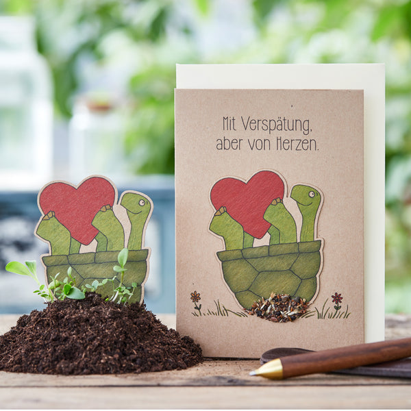 Grußkarte mit gezeichneten Schildkröten, die ein rotes Herz halten, daneben ein passender Pflanzenstecker in Erde; Text auf der Karte: "Mit Verspätung, aber von Herzen."