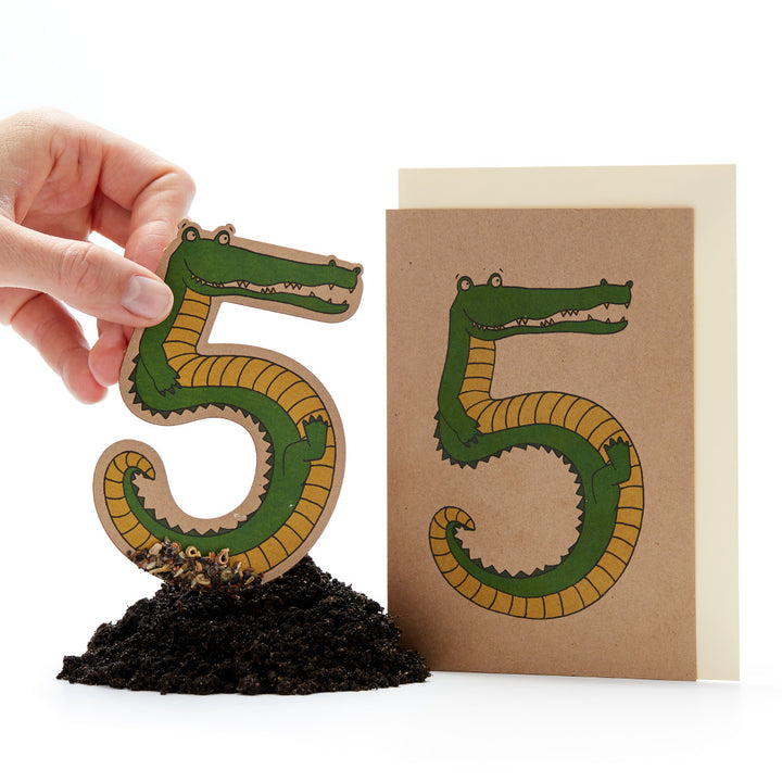 Hand hält eine ausgeschnittene Papierfigur in Form einer "5", die wie ein Krokodil aussieht, neben einer Karte mit dem gleichen Design auf Erde.