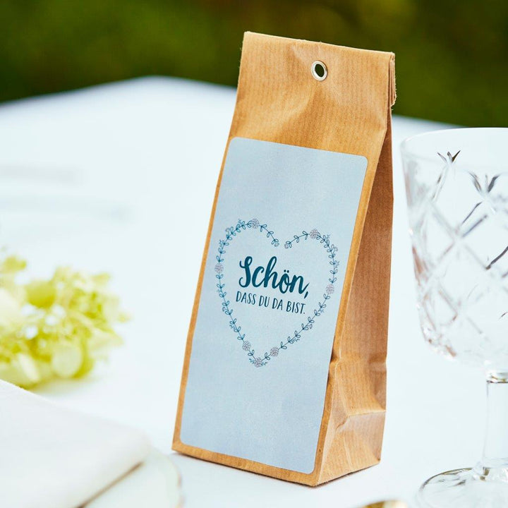 Braune Papiertüte mit der Aufschrift "Schön, dass du da bist" auf einem Tisch neben Weinglas und Trauben.