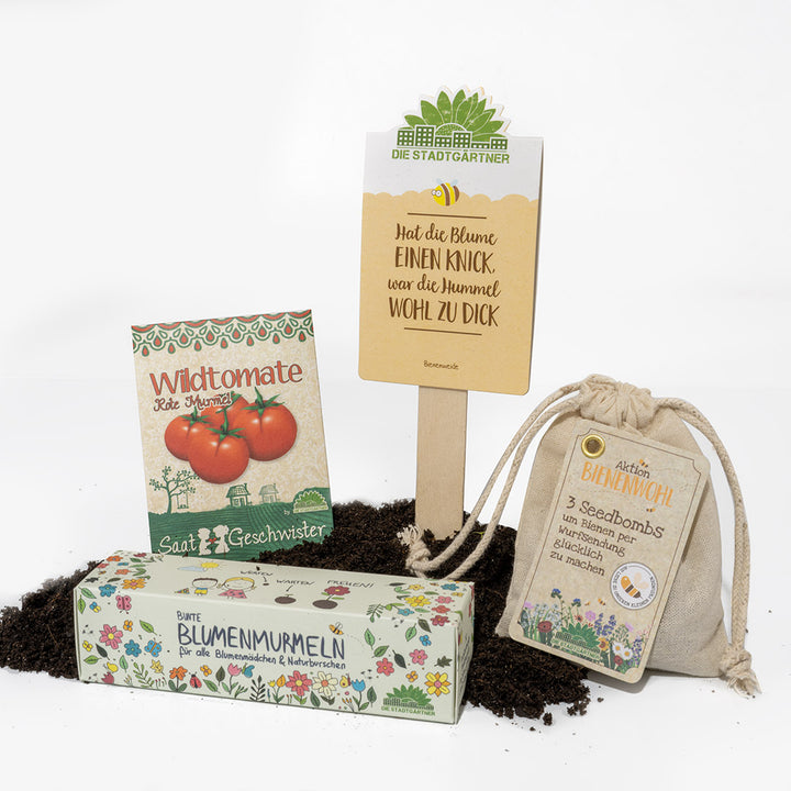 Gartenprodukte auf Erde, inklusive einer Tüte mit 'Wildtomate' Samen, 'Blumenmurmeln', 'Bienenwohl' Säckchen und einem Schild 'Die Stadtgärtner'.